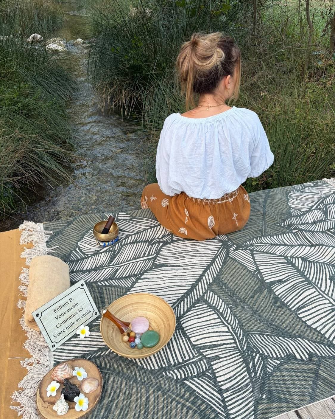 Annabelle assise au bord d'une rivière, bol chantant et pierres posés près d'elle